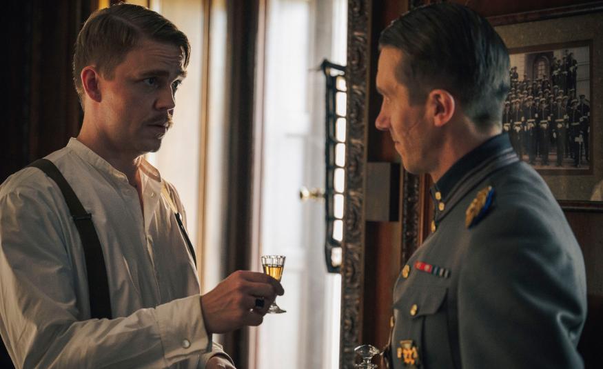 Kuvassa on vasemmalla Aku Sipola valkoisessa paidassa kohottamassa lasia sotilaspuvussa olevalla Jussi Vataselle, joka on osittain selin. Taustalla on iso ikkuna ja kuva sävy on tummahko. 