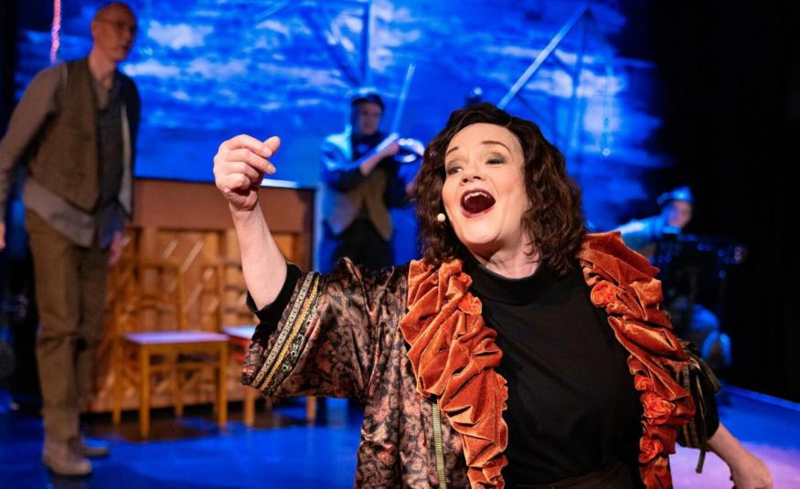 Kuvassa on Lise Holmberg keskellä laulamassa mustassa mekossä ja ruskehtavassa rimpsujakussa. Taustalla on  vasemmalla mies ruskehtavissa vaatteissa ja takana on soittaja.