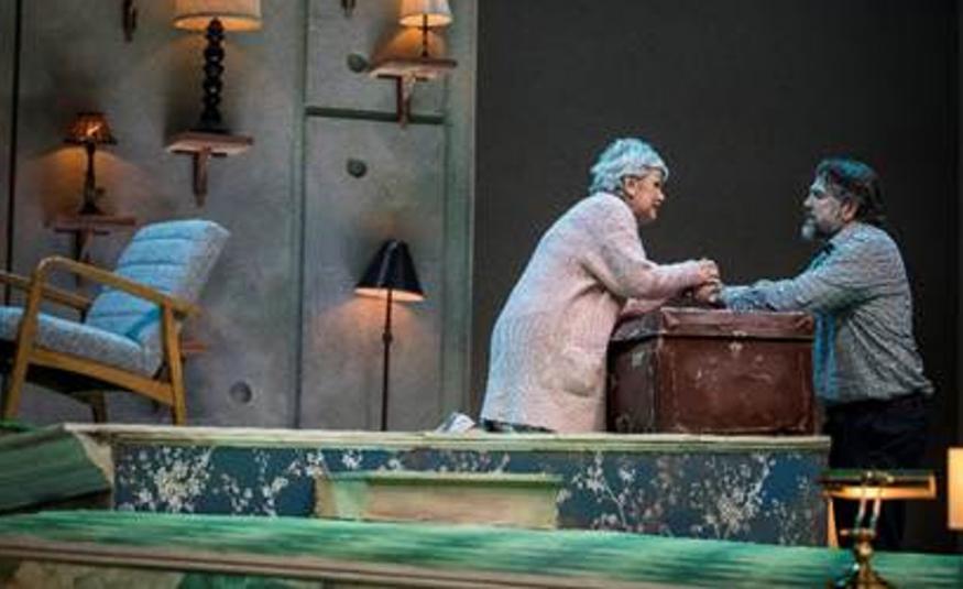 Kuvassa on huone ja vasemmalla nojatuoli sekä seinällä lamppuja. Oikealla on ruskean tavara-arkun ääressä seisomassa kumarassa naisen ja toisella puolella on mies.