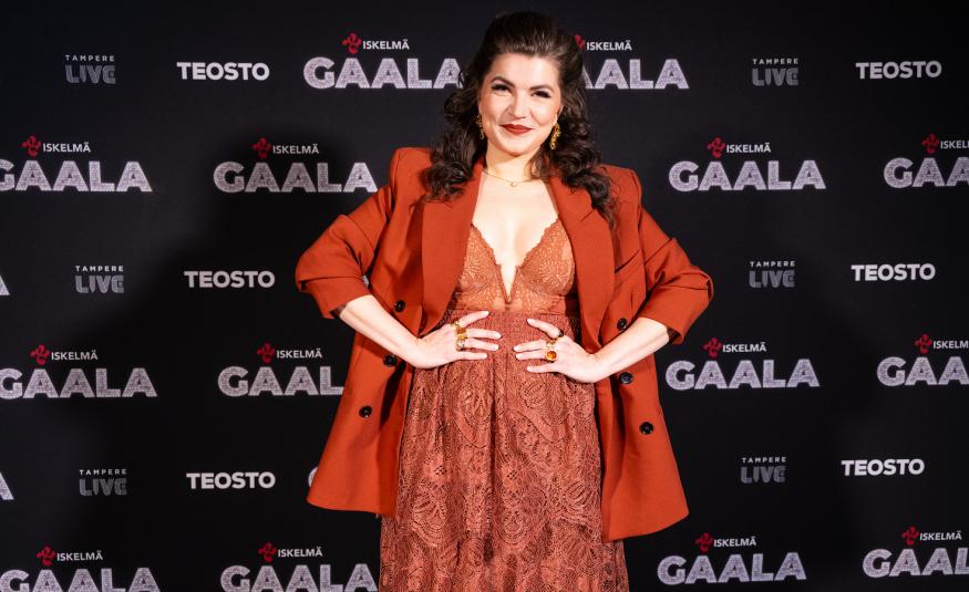 Kuvassa on Suvi Teräsniska tumman oranssin ruskeassa mekossa ja jakussa.  Hänellä on kädet lanteillaan. Taustalla on tummapohjaisella seinällä ja siinä lukee moneen kertaan Gaala ja Teosto.