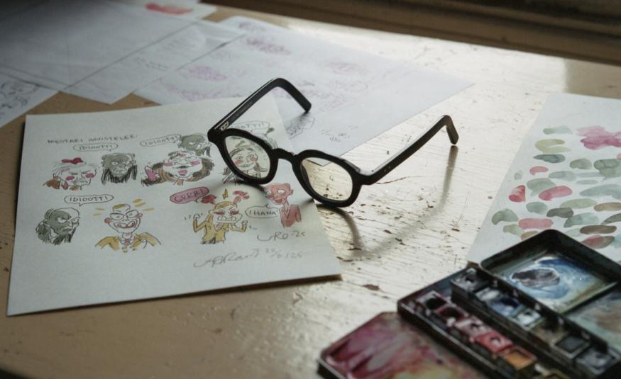 Kuvassa on pilapiirroskuvia pöydällä valkoisella paperilla ja niiden päällä silmälasit. Oikeassa reunassa on värejä.