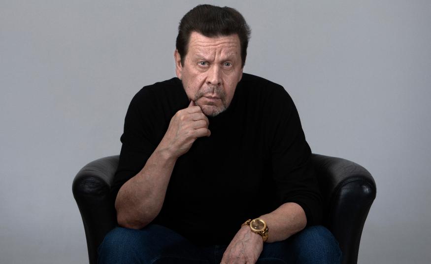 Kuvassa on Pate Mustajärvi istumassa mustassa nojatuolissa vähän kumartuneena teenpäin ja hän nojaa oikealla kädellään leukaansa. Hänellä on musta pusero päällään. Tausta on harmaa.