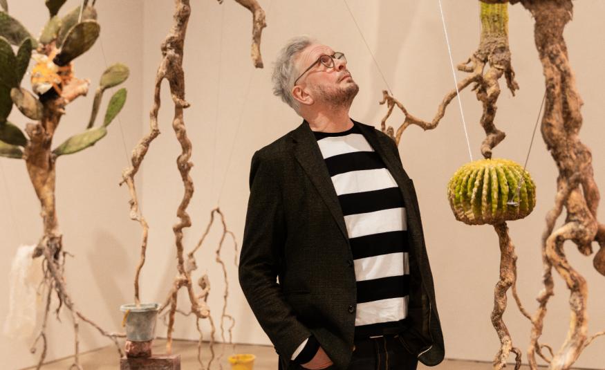 Kuvassa on keskellä Roland Persson seisomassa puolivartalokuvassa. Hänellä on musta takki ja musta-valkoinen paksuraidallinen pusero päällään. Hän katsoo ylöspäin. Taustalla on roikkuvia juurista tehtyjä taideteoksia ja seinät ovat vaalean ruskeat.