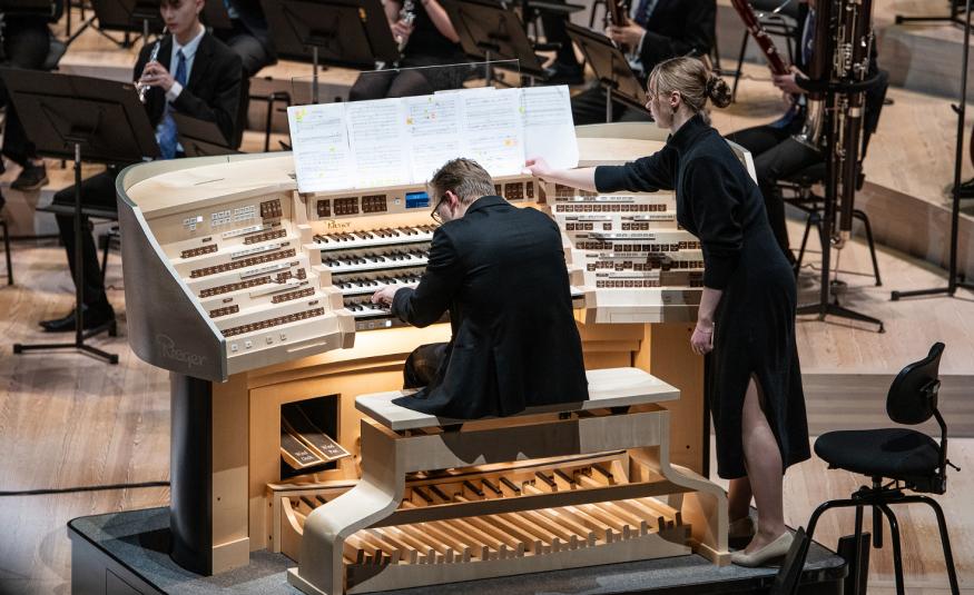 Kuvassa on keskellä selin soittamassa urkuja Aleksanteri Wallius ja urut ovat leveästi orkesterin edessä. Orkesteria näkyy hieman. Urkurin vieressä seisoo nainen, joka kääntää nuottia urkurille.