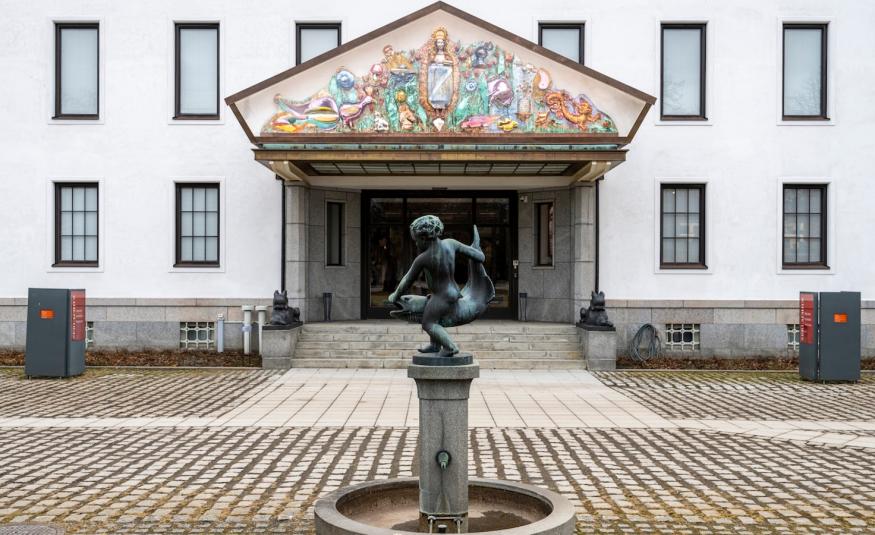 Kuvassa on valkoinen rakennus jossa on kapeita ikkunoita kahdessa kerroksessa. Siinä on portaikko ja isot ovet ja niiden päällä iso kolmio. Siinä on reliefimäisesti palstellivärisiä hahmoja ja esineitä.