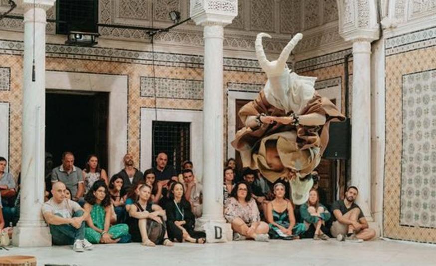 Kuvassa on joukko ihmisiä istumassa rakennuksen valkoisella lattialla ryhmänä ja edessä on kolme valkoista, itämaista pylvästä. taustalla näkyy seinää, jossa on valkoinen, avoin oviaukko ja vieressä kaksi pienempää aukkoa. Seinässä on keltakuvioista mosaiikkia ja sinikuvioista ornamenttia. 