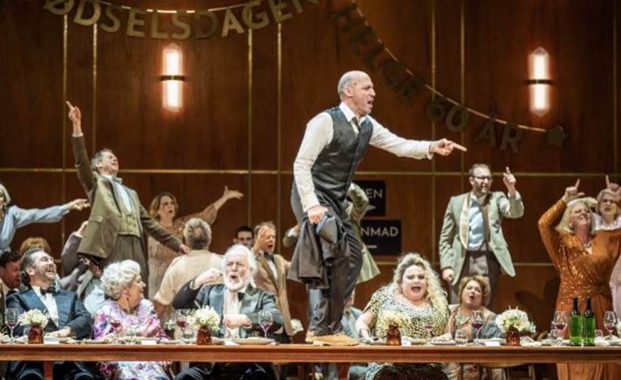 Kuvassa on ihmisiä puvuissa juhlimassa pitkän pöydän ääressä ja takana on juhlivia häenkilöitä kädet levitettynä. Keskellä on mies seisomassa pöydällä sivuttain ja hänellä on harmaa liivi ja housut sekä valkoinen paita. Taustalla on ruskea seinä.