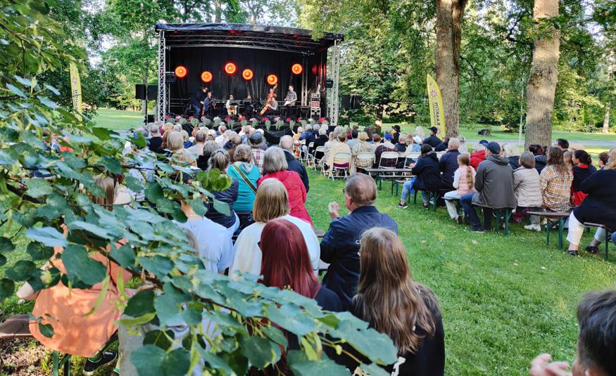 Kuvassa on puiston vihreitä puita ja oksia. Taustalla on esiintymislava ja siinä orkesteri soittamassa. Lavan edestä taakse päin on yleisöä istumassa tuoleilla.