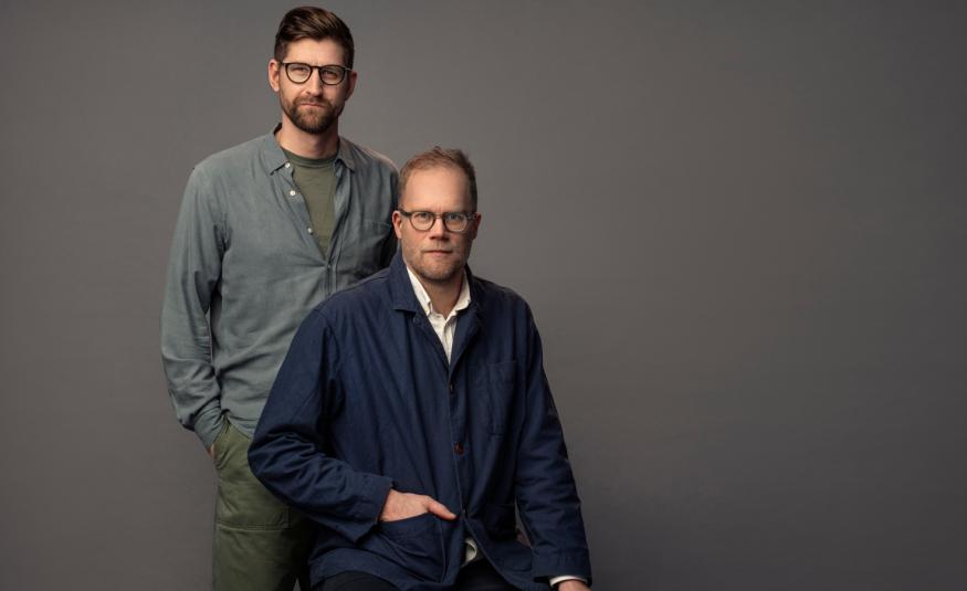 Kuvassa ovat Lars Berge seisomassa harmaassa puserossa Axel Gords Humlesjön takana ja Humlesjö istuu. Hänellä on musta pusero ja pitkät housut. Tausta on tumman harmaa.