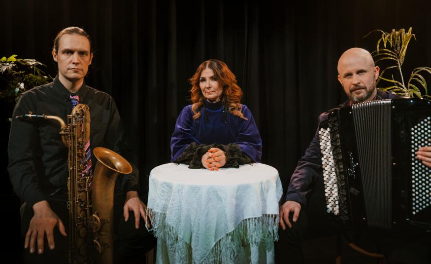 Kuvassa ovat valkoisen liinan peittämän pienen pöydän ääressä vasemmalla Joakim Berghäll ruskehtava saksofoni, keskellä Irina mustassa puserossa ja oikealla istumassa harmonikka sylissään.