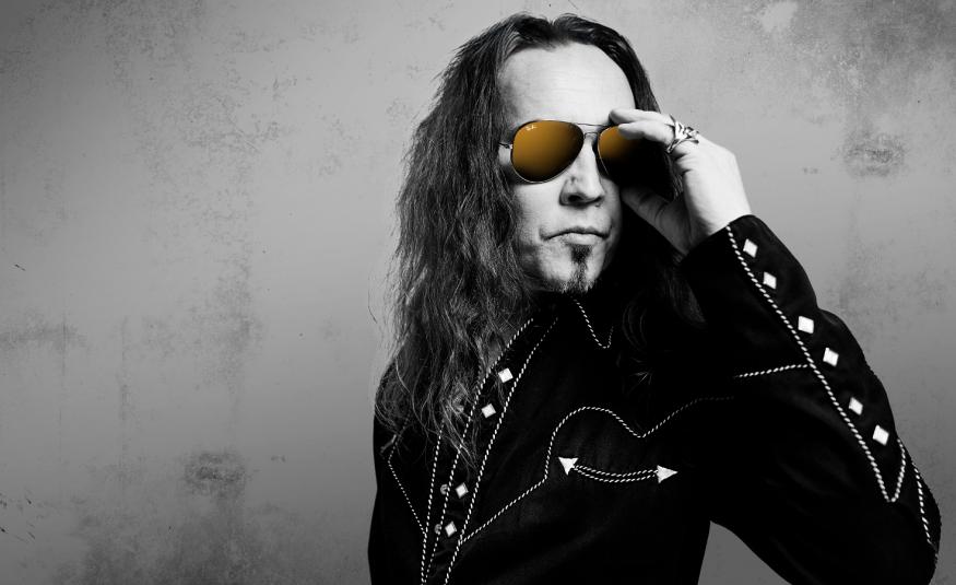 Kuvassa on Jukka ahola puolivartalokuvassa ja hänellä on musta takki päällään. Siinä on nappeja hihassa ja Ahola pitää vasemmalla kädellään aurinkolaseista kiinni. Kuva on musta-valkoinen tausta on harmaa.