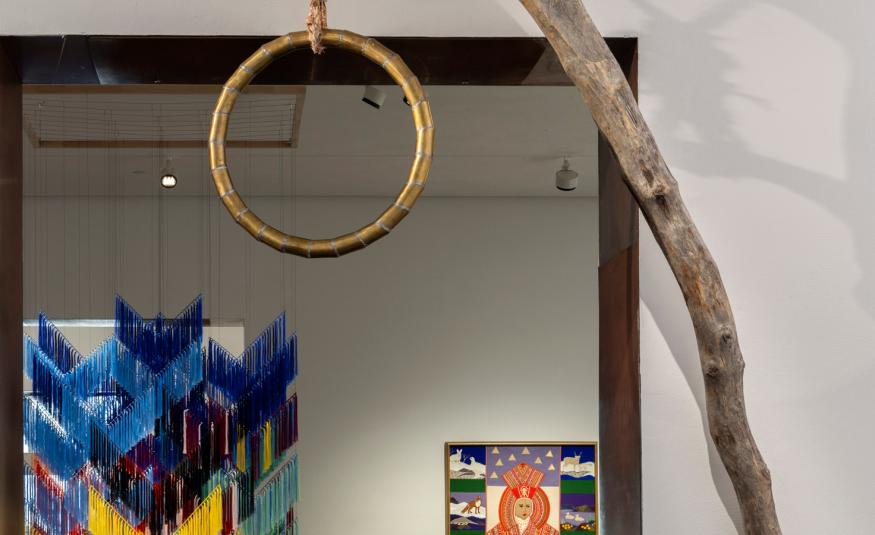 Kuvassa on etualalla kapeaa puunrunkoa ja siitä roikkuu puinen, pyöreä hirttosilmukka. Taustalla näkyy roikkuva tekstiilityö, jossa on Lapin värejä ja näyttelysalia.