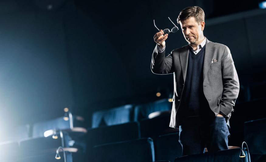 Kuvassa on oikealla Klaus Härö seisomassa ja puhumassa lavalla, jossa on taustalla istuinrivejä. Hänellä on harmaa takki ja tumman sininen liivi. Hän pitää silmälaseja oikeassa kädessään ylhäällä. Tausta on tumman sininen ja vasemmalle heijastuu valoa.