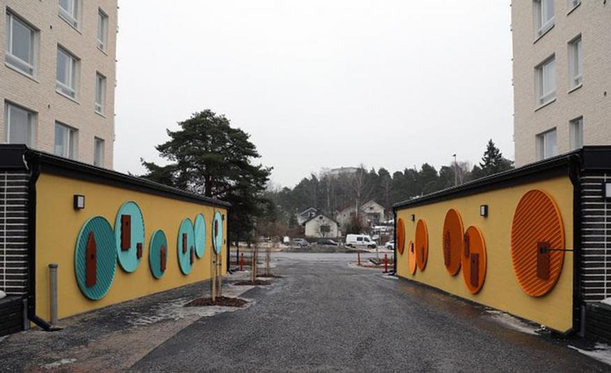 Kuvassa on molemmissa reunoissa vaalean ruskeat kerrostalot. Vasemmalla on pitkä alarakennus ja siinä ruskehtavalla pohjalla on isoja pyöreitä turkooseja alustoja, joilla on tummia yksityiskohtia. Toisella puolella sisääntulotie ja sen toisella puolella vastaava alarakennus ja siinä punaiset pyöreät koristeet. 