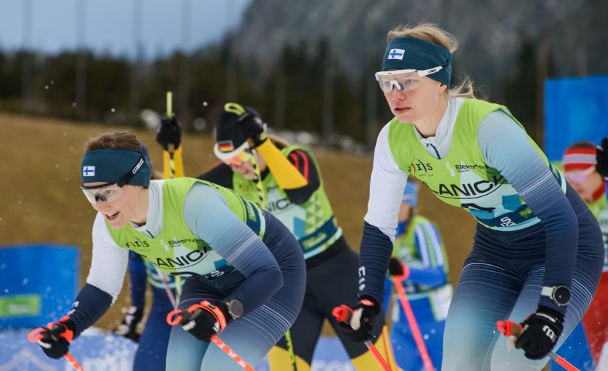 Kuvassa on kolme naishiihtäjää lähdössä kilpailemaan. Heillä on Suomen sini-valkoiset edustusasut ja pipot, joissa on Suomen lippu. 