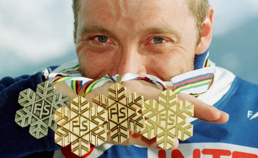 Kuvassa on taustalla Mika Myllylän kasvot ja hän pitää puoliksi kasvojen edessä neljä mitalia, joissa on havukuviota ja keskellä kirjaimet FS. taustalla vaalean sinistä pilveä.