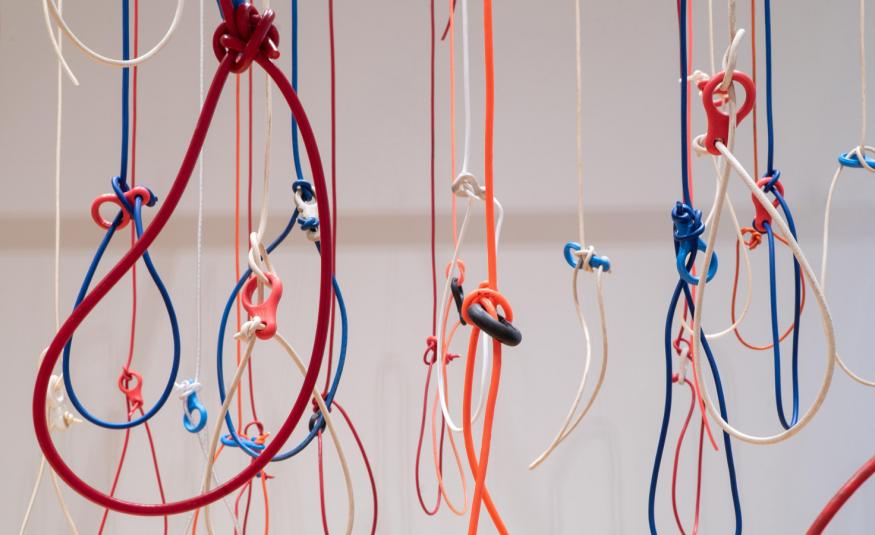 Kuvassa on roikkumassa erivärisiä naruja ja niissä on solmuja. Joihinkin on solmittu iso silmukka. Tausta on harmaa.