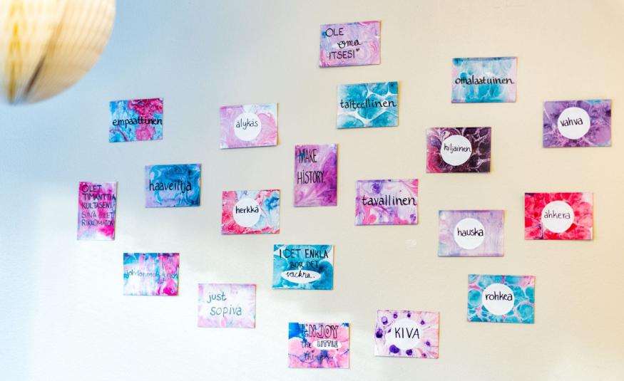 Kuvassa on vaalean sinisellä ja vaalean keltaisella pohjalla lappuja seinällä ja niiden värit ovat puna-sini-valkoisia.  Niissä on tekstejä kuten kiva, rohkea, herkkä jne.