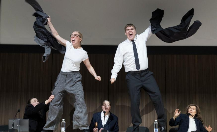 Kuvassa on nuori nainen vasemmalla ja hän heiluttaa takkiaan ja hänellä on valkoinen t-paita ja harmaat, löysät housut päällää, Oikealla on nuori mies, jolla on valkoinen paita jamusta kravatti sekä mustat housut päällään. Taustalla alhaalla on kolme henkilöä pöydän ääressä mustissa takeissa.