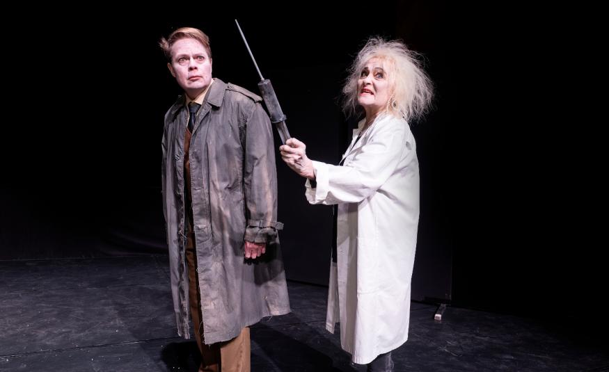 Kuvassa on mies ruskeassa päällystakissa ja oikealla nainen vakoisessa päällystakissa. Tausta on tumma.