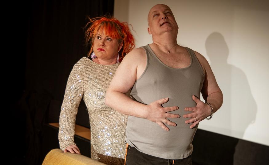 Kuvassa on vasemmalla vaaleassa glitterpuserossa Mira Lukkari seisomassa ja nojaamassa sohvan selkämykseen oikealla kädellään.  Hänen edessään on Jari Lukkari seisomassa harmaassa hihattomassa t-paidassa ja taputtaa oikealla kädellään vatsaansa. 