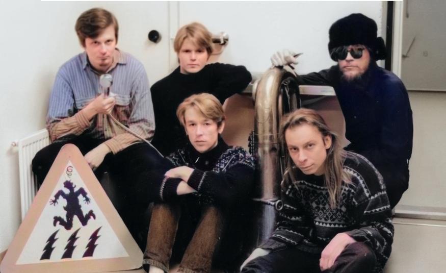 Kuvassa on viisi nuorta miestä, joista kolme istuu takarivissä ja heistä yhdellä on vaalean sininen pusero ja muilla mustat puserot. Edessä istuu kaksi miestä ja vasemmalla heidän vieressään on kolmioliikkennemerkki jossa on pomppivan henkilön kuva. Kuva on osa kirjan kannesta.