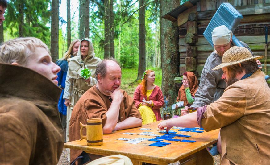 Kuvassa on metsässä olevan harmaan hirsimökin vieressä pöydän ääressä vastakkain kaksi miestä ja pöydällä on sinisiä kortteja, joita katsovat. Pöydän päässä seiroo nainen harmaassa huivissa ja vasemmassa kulmassa ruskeassa takissa nuori mies. Taustalla näkyy ihmisiä keskiaikaisissa vaatteissa.