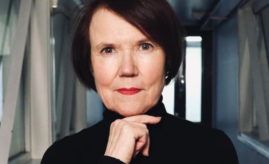 Kuvassa on Rosa Liksom puolivartalokuvassa mustassa poolopuserossa ja hänellä on oikea käsi leuan alla. Hänellä on tummat, punertavat puolipitkät hiukset. Taustalla on harmaata käytävää.