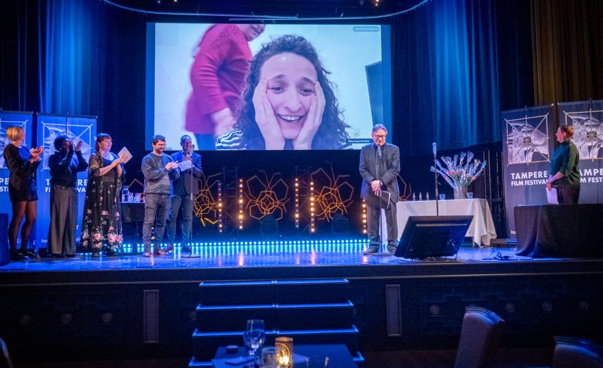 Kuvassa on sinisävyinen näyttämö ja vasemmalla seisoo viisi henkilöä taputtamassa. Oikealla seisoo mies, joka puhuu. Taustalla näkyy ylhäällä valkokangas ja siinä on naisen kasvot. Hän pitää käsiään poskillaan.