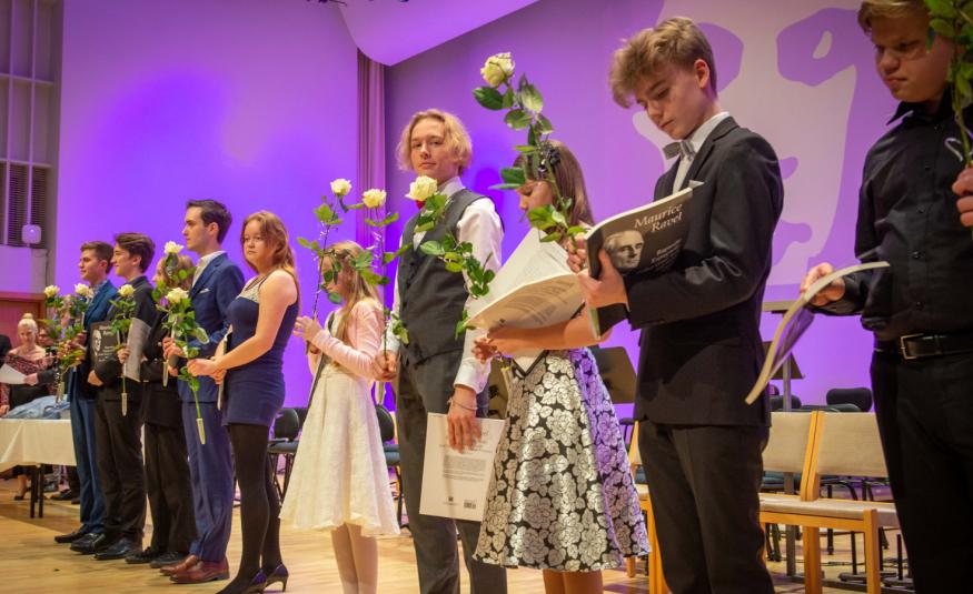 Kuvassa on viistosti seisomassa näyttämöllä sekä tyttöjä että poikia. Heillä on kädessään ruusu ja kunniakirja. Taustalla on violettia valoa.