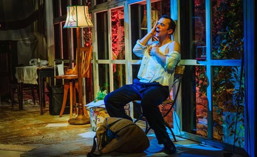 Kuvassa on Ilja Peltonen kalustetussa huoneessa, jossa Ilja Peltonen istuu tuolilla ja nojaa taaksepäin.  Hänellä on vaalean sininen paita ja siniset housut päällään. Huoneessa on pieni pöytä ja iso jalkalamppu sekä takana isot ikkunat. 