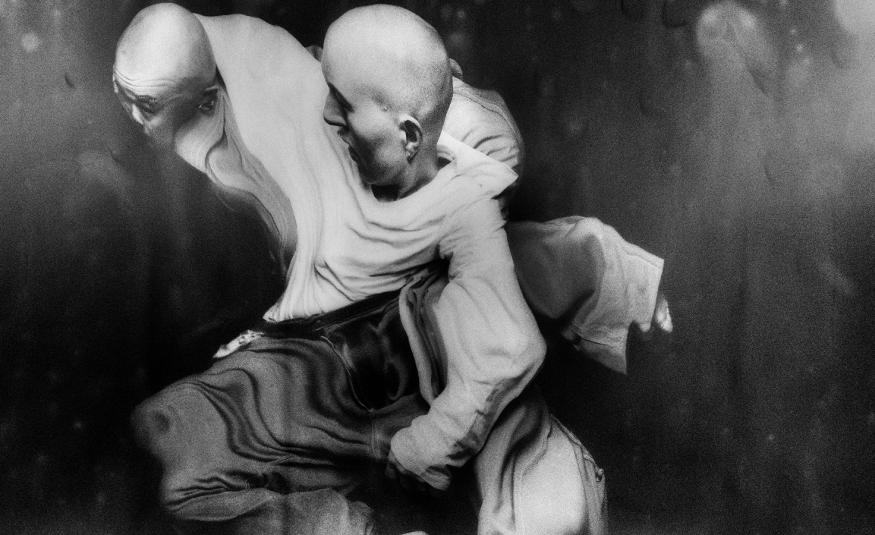 Kuvassa on kaksi miestanssijaa, jotka ovat kietoutuneitä toisiinsa vaalealla paidalla. Heillä on tummat housut. Kuva on musta-valkoinen.