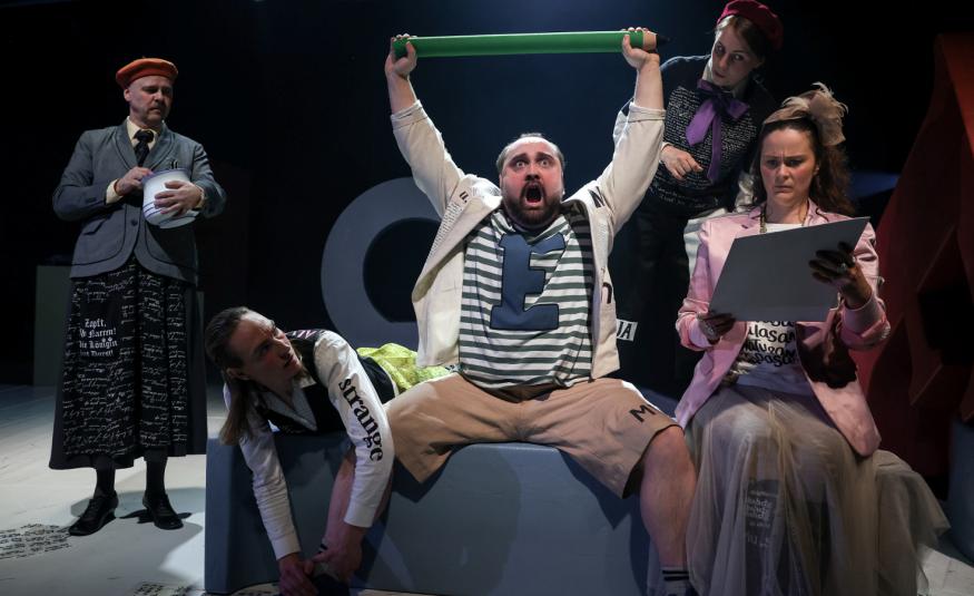 Kuvassa on keskellä Sami Sainio, joka istuu sohvalla ja pitää vihreää keppiä ylhäällä käsissään.  Vieressä istuu nainen vaaleassa takissa. Hänellä on kansia kädessään. Taustalla seisoo harmaassa takissa nainen.