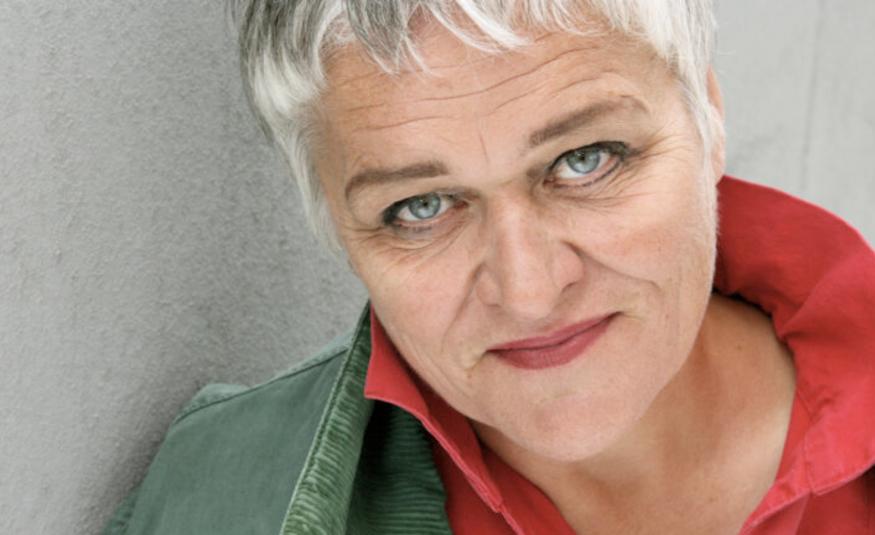 Kuvassa on Pirkko Saision kasvokuva ja hänellä näkyy kuvassa harmaata takkia ja punaisia kauluksia. Hänellä on vaalean harmaat hiukset.