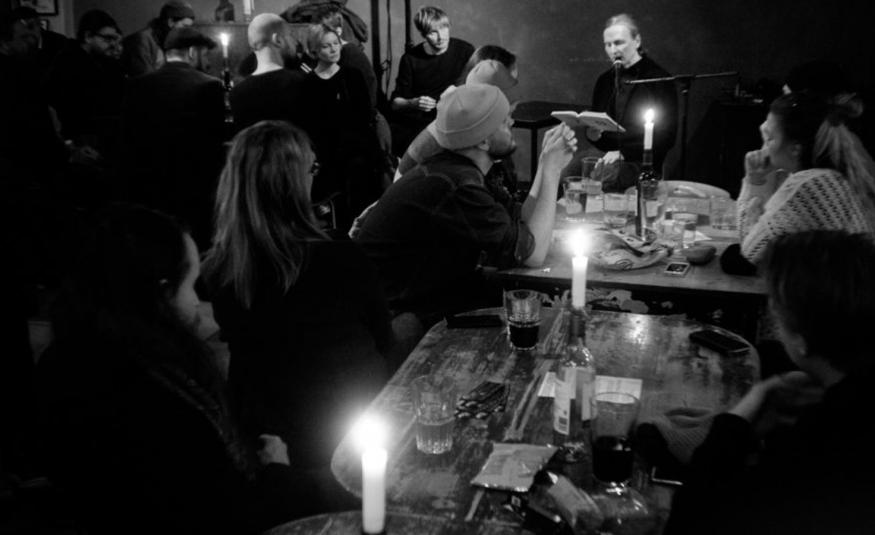 Kuvassa on huone täynnä ihmisiä. Oikealla on pitkä pöytä ja sen äääressä on ihmisiä juomassa ja keskustelemassa. Pöydässä on kynttilöitä. Vasemmalla on ihmisiä seisomassa. Kuva on musta-valkoinen.