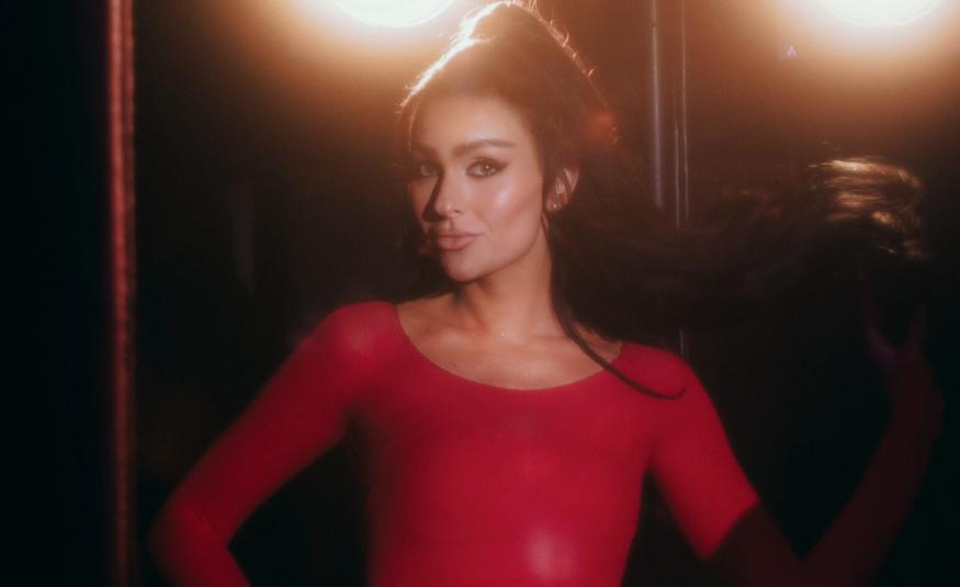 Kuvassa on keskellä Sara Siipola puolivartalokuvassa. Hänellä on punainen mekko päällään ja hänellä on tummat hiukset nutturalla. Taustalla tumman ruskeaa ja kaksi ylävaloa.