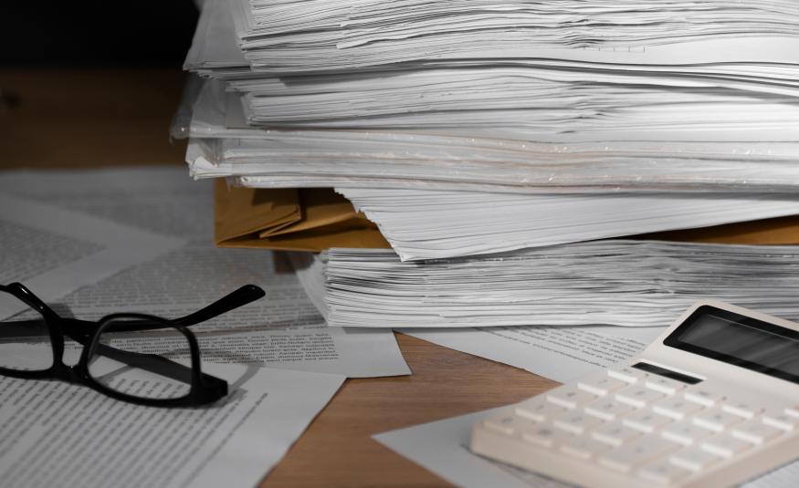 Kuvassa on iso pino valkoisia papereita ja vasemmalla on silmälasit ja oikealla edessä on valkoinen laskin. 