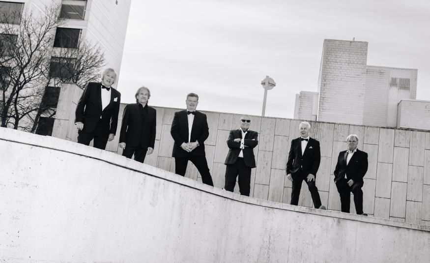 Kuvassa on alaspäin viettävän kaiteen vierellä kuusi mieslaulajaa seisomassa mustissa puvuissa.  Taustalla on vaaleaa seinämää ja vasemmalla kerrostalon julkisivua ikkunoineen. Kuva on musta-valkoinen.