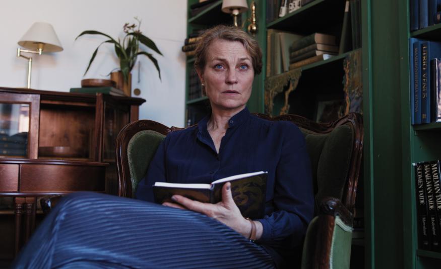 Kuvassa on Jonna Järnefelt istumassa nojatuolissa ja hänellä on kädessään muistikirja kädessään. Takana on ruskea matala kirjahylly ja oikealla on iso tumman ruskea kirjahylly.