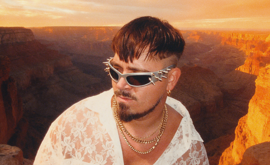 Kuvassa on Käärijä keskellä alempana ja hänellä on valkoinen paita päällään ja aurinkolasit. Taustalla on ilta-auringon valossa olevaa Grand Canonia.