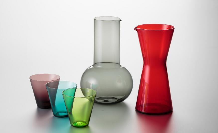 Kuvassa on vasemmalla kolme eri väristä lasia ja keskellä harmaa iso alhaalta pyöristetty maljakko. Oikealla on punainen kaatomaljakko. Tausta on vaalean harmaa.