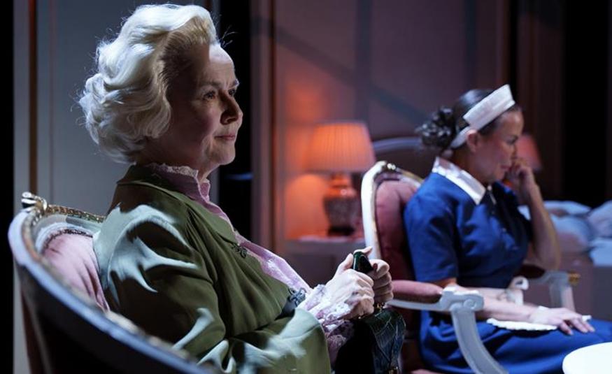 Kuvassa on etualalla Paula Siimes istumassa nojatuolissa vinottain ja hän pitää kävelykepistä kiinni edessään. Taustalla näkyy Maria Kuusiluoma istumassa valkoinen hilkka päässään.