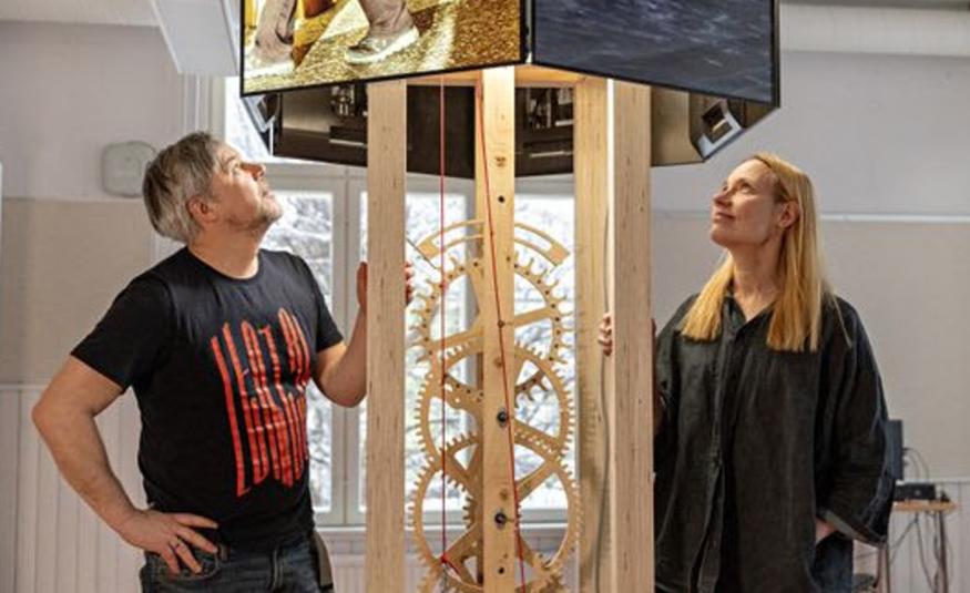 Kuvassa on vasemmalla Oliver Kochta-Kalleinen katsomassa ylöspäin puisen kellokoneiston yläpuolella olevaa neliötä ja sen koneistoa. Oikealla on Tellervo Kalleinen seisomassa ja katsomassa myös ylöspäin. Hänellä on musta leveä mekko. Taustalla on vaalealla seinällä ikkunoita.