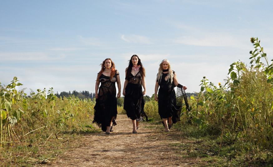 Kuvassa ovat Rodeon kolme jäsentä kävelyllä hiekkatiellä ja vierellä on korkeita kasveja pellolla. Heillä tummat pitkät mekot päällään. Taustalla on sinistä taivasta.