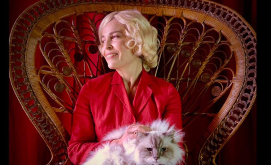 Kuvassa on Irina Björklund istumassa isossa korituolissa ja hänellä on valkoinen kissa sylissään. Björklundilla on vaaleat hiukset ja punainen mekko päällään. Takaseinä on punainen.