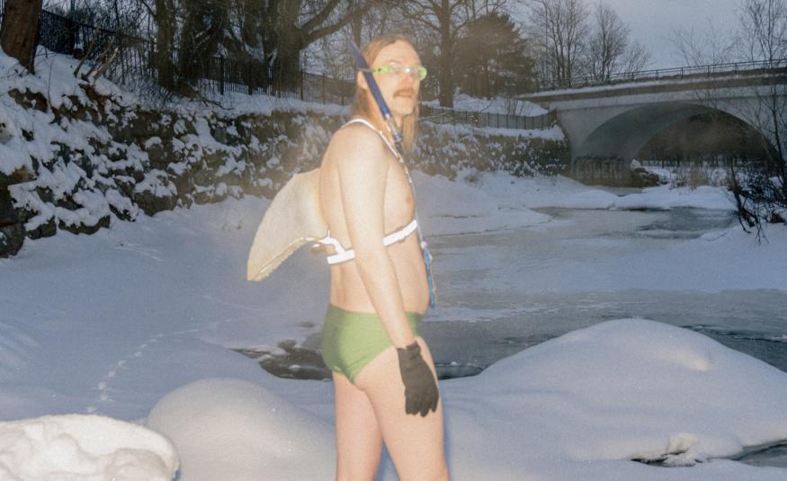 Kuvassa on Stig menossa lumisessa maisemassa sillan ali vitaavaan veteen uimaan. Hänella on valkoiset siivet selässään ja vaalean vihreät uimahousut sekä mustat hansikkaat.