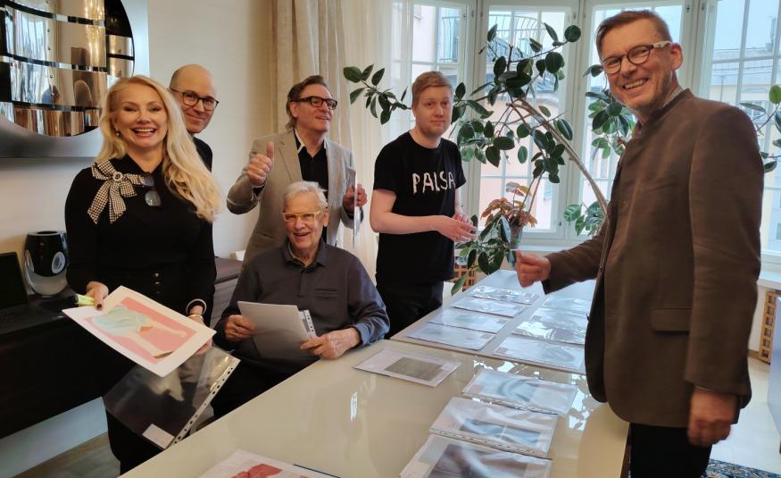 Kuvassa ovat suuren pöydän ääressä seisomassa on vasemmalla viisi henkilöä ja edessä oikealla seisoo yksi mies. Pöydällä on taideteosten luonnoksia A4 koossa.