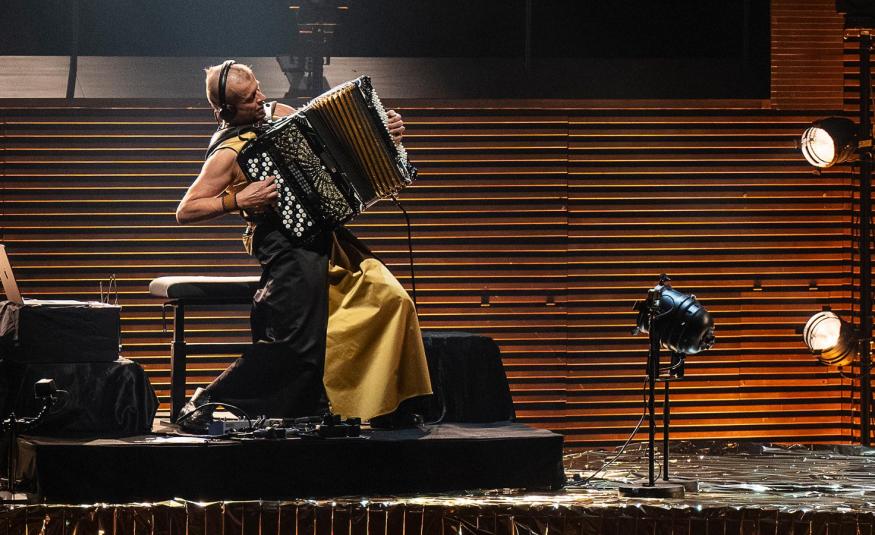 Kuvassa on vasemmalla jalustalla sivuttain Kimmo Pohjonen keltaisissa leveissä housuissa soittamassa harmonikkaa. Takana on ruskeaa vaakasuoraa rimaa.