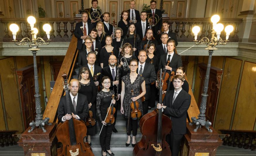 Kuvassa on Vaasan kaupunginorkesterin jäsenet seisomassa leveillä portailla ja taustalla sekä sivuilla on keskiruskeaa panelointi. Ylhäällä on molemmin puolin kolmen lampun valaisimet. 