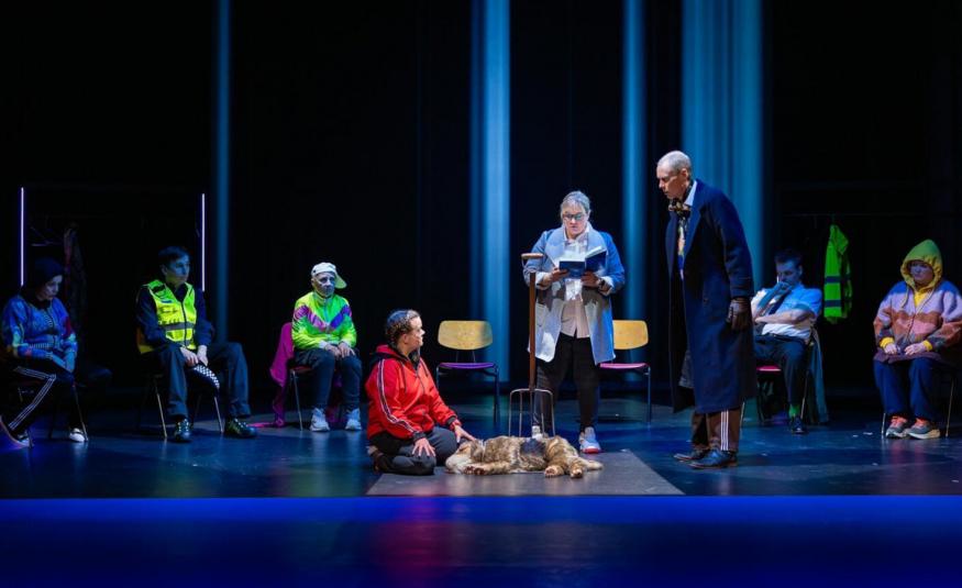 Kuvassa on tummalla näyttämöllä edessä mies punaisessa pusakassa polvillaan ja hänen edessään on vaalean ruskea koira. Hänen vieressään seisoo nainen lukemassa kirjasta tekstiä ja hänellä on vaalean sininen jakku. Hänen vierellään oikealla seisoo mies tummassa takissa. Vasemmalla istuu kolme henkilöä ja oikealla kaksi pusakoissa.